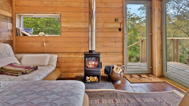 A cozy cabin interior with a wood-burning stove, a sofa, and a window overlooking greenery. - Beautiful  villa for 2 guests (Dingwall)