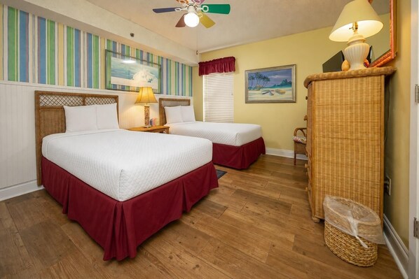 A room with two beds, a ceiling fan, a lamp, a wicker cabinet, and a painting on the wall.
