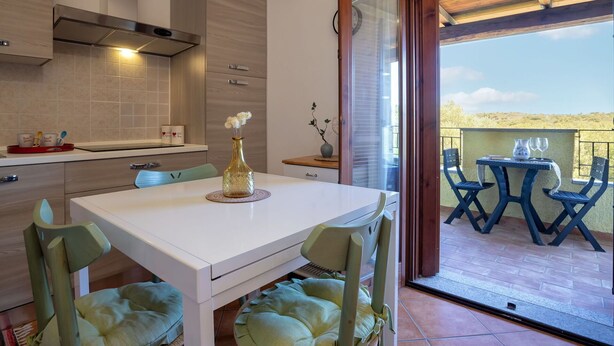 A modern kitchen with a white countertop, a stainless steel range hood, and a dining area with a white table and green upholstered chairs. - Nice apartment for 4 people with A/C, TV, balcony and panoramic view (Olbia)