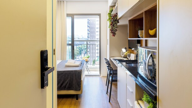 Une chambre d’hôtel compacte comprenant un lit, une petite kitchenette, un coin repas avec une table et des chaises, et une vue sur la ville grâce à une grande fenêtre.