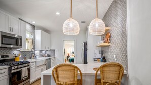 A modern kitchen with white cabinets, a stainless steel stove, and a central island with wicker chairs. - Sandy Toes Retreat | Charming, Pool Access (Galveston)