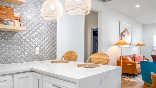 A modern kitchen with white cabinets, a marble island, and a backsplash with a shell pattern. There are wicker chairs and a blue armchair in the dining area. - Sandy Toes Retreat | Charming, Pool Access (Galveston)