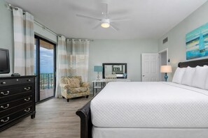 Habitación de hotel con cama, cómoda, silla, escritorio con espejo, ventilador de techo y un cuadro en la pared.