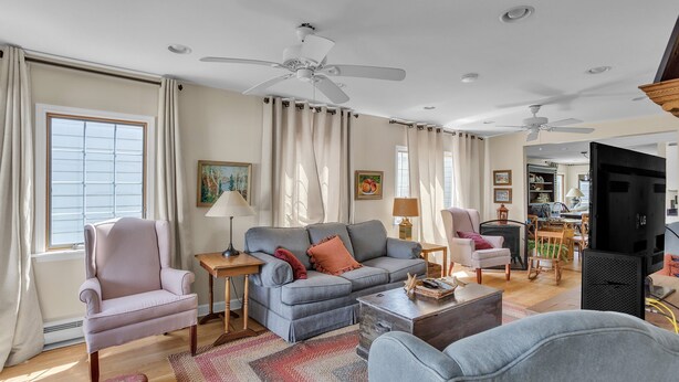 A spacious living room with a grey sofa, a pink armchair, and a wooden coffee table. There are ceiling fans, a TV, and a dining area visible. - Home Port - Spacious Retreat In Prime Location 4 Bedroom Home (Davis)