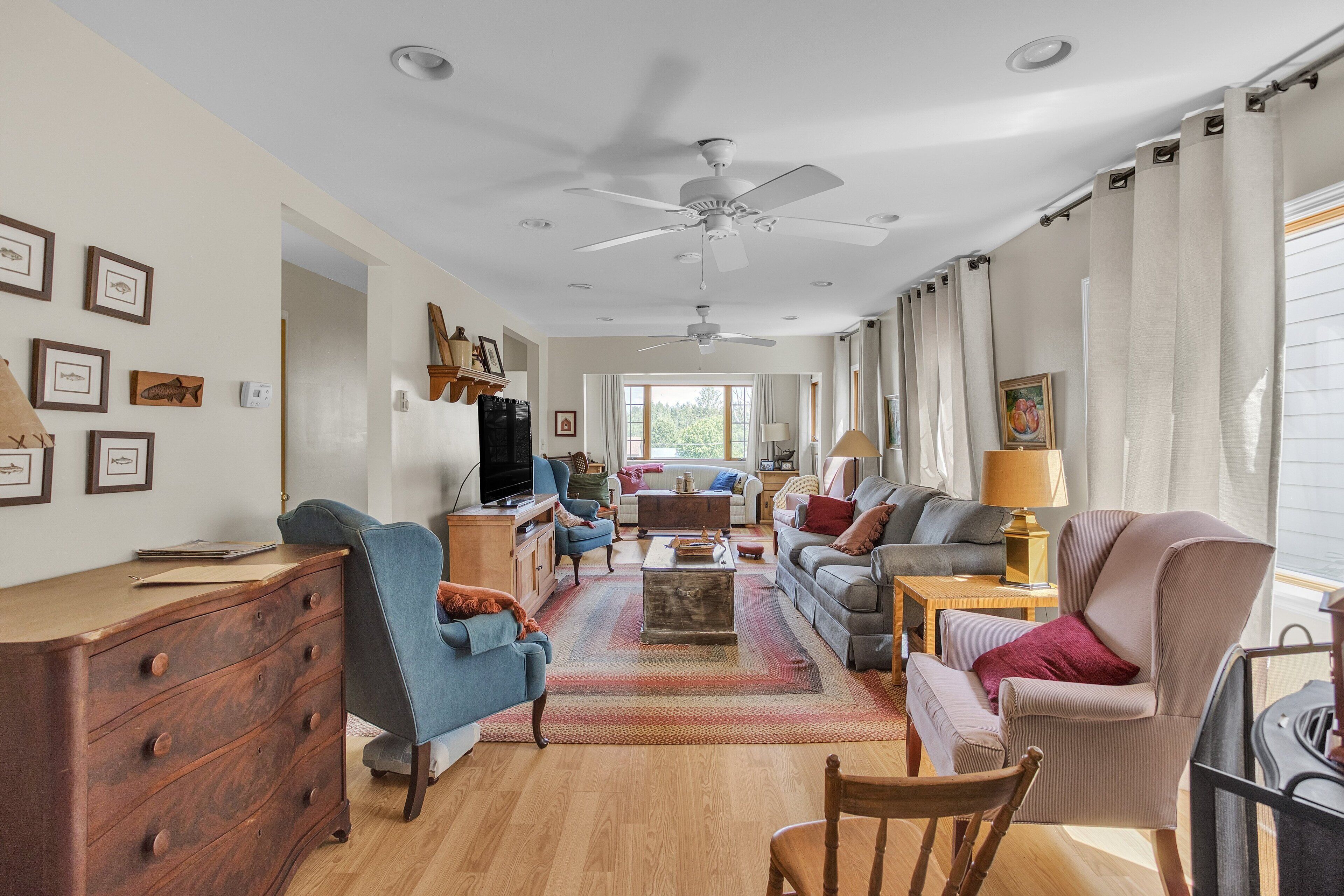 A spacious living room with a ceiling fan, wooden furniture, and a large window with curtains.