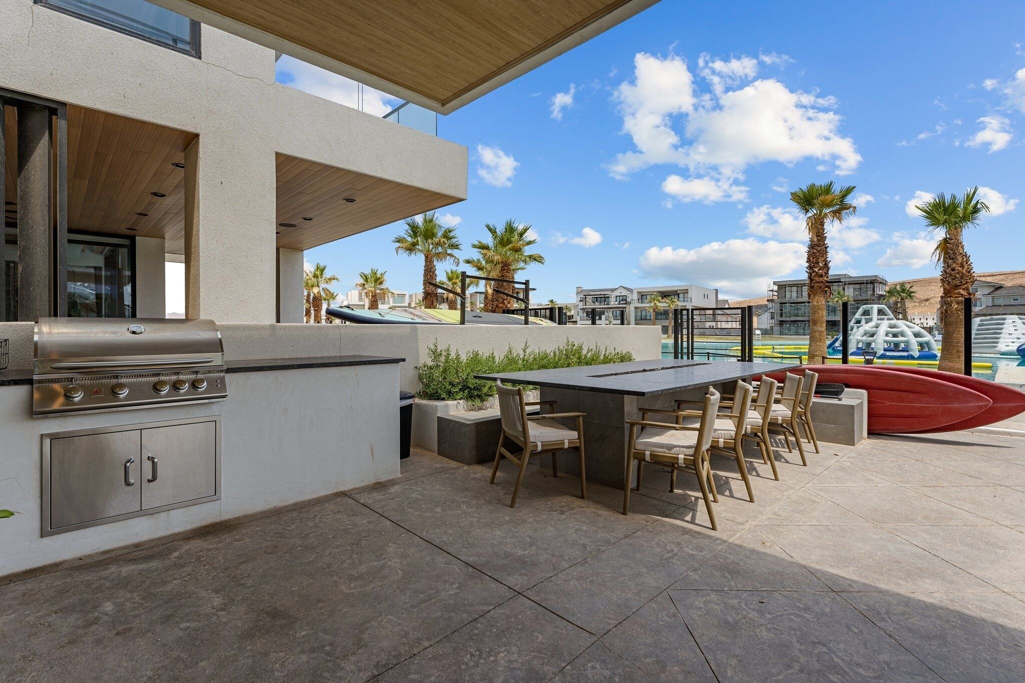 An outdoor kitchen with a grill, dining table, and chairs, surrounded by palm trees and modern buildings.