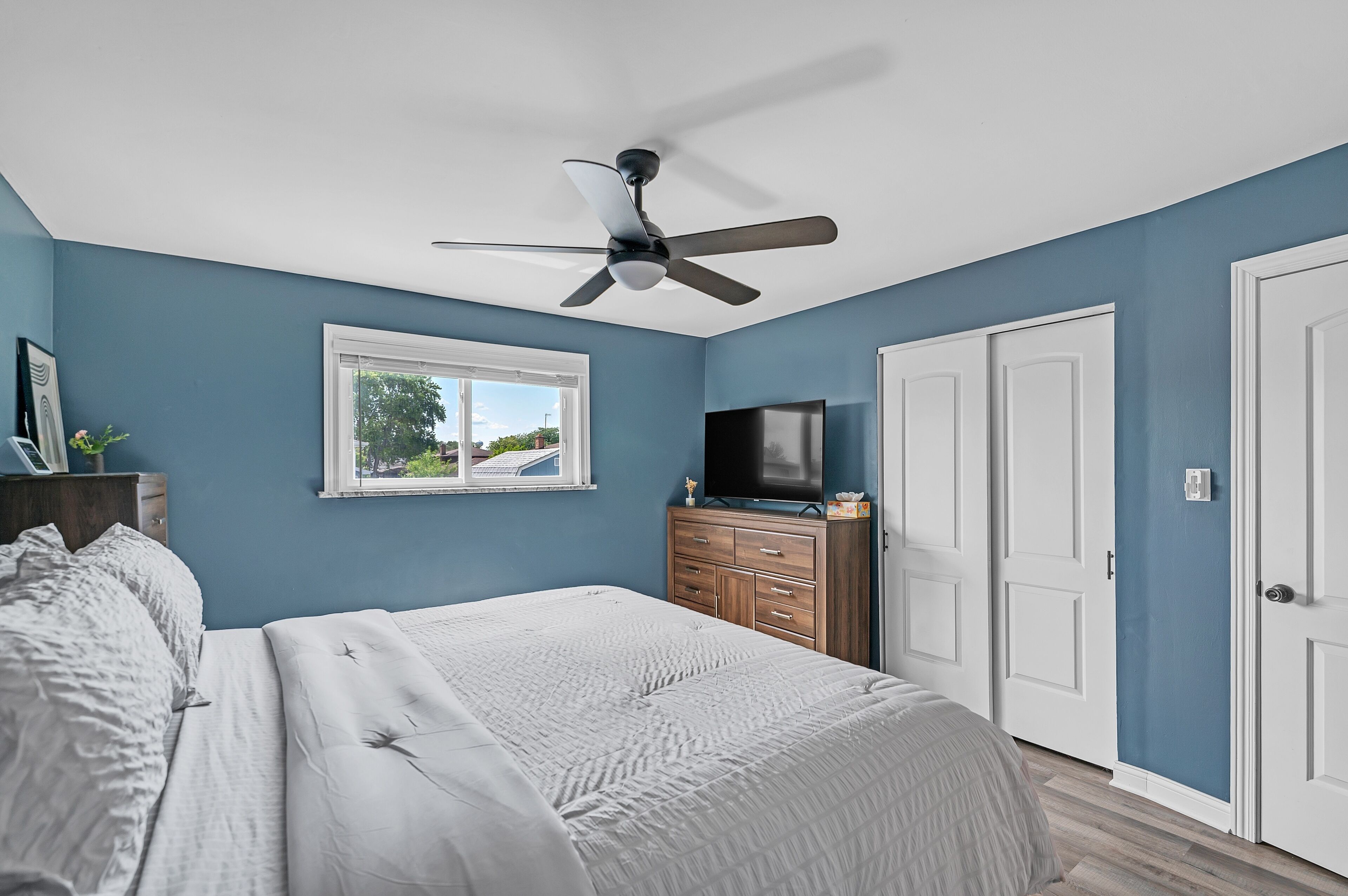 A bedroom with a bed, a ceiling fan, a TV, a window, and a door.