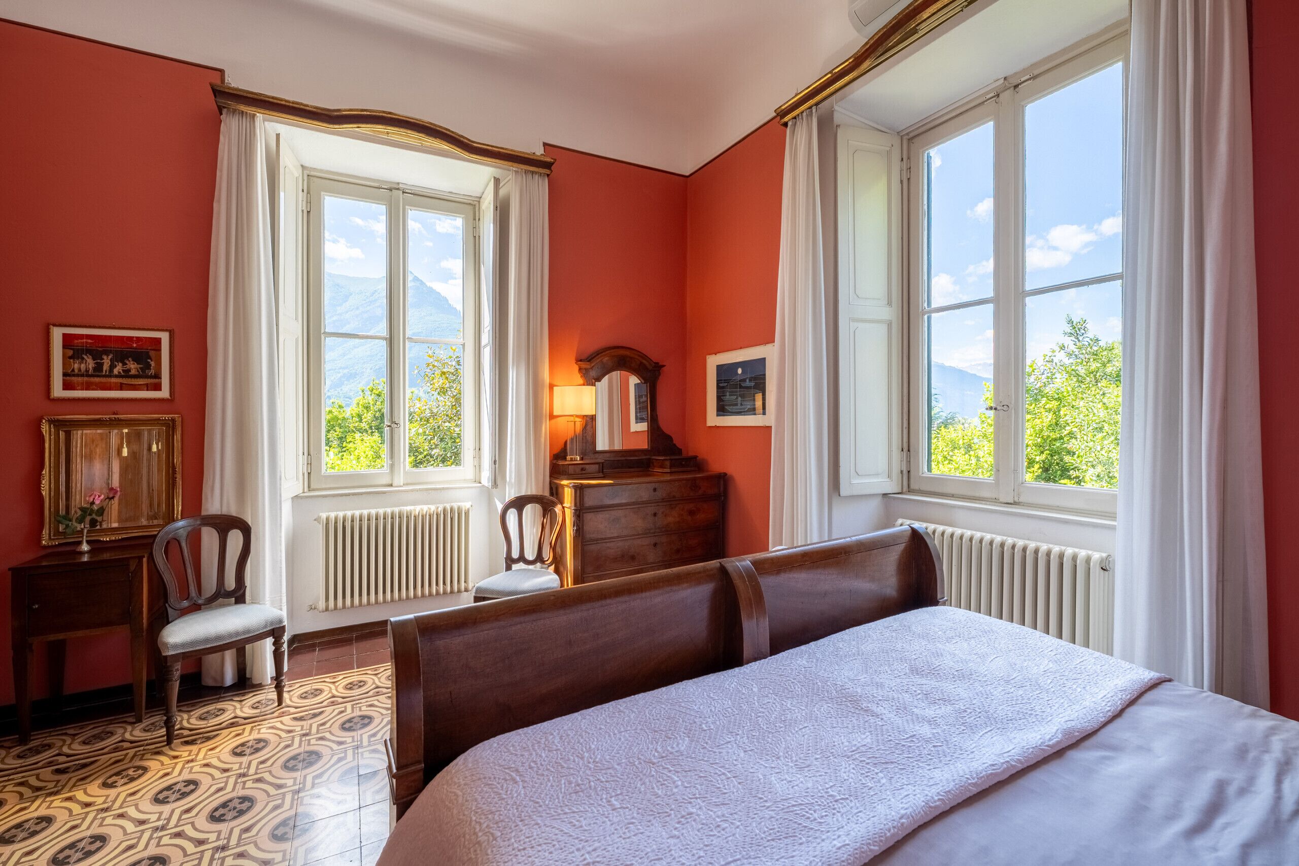 A bedroom with a large bed, two windows with curtains, a dresser, a chair, and a radiator.