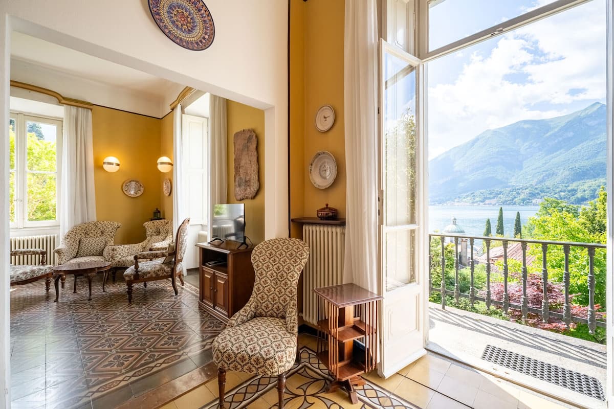 A spacious living room with patterned furniture, a view of a lake, and a balcony with a railing.