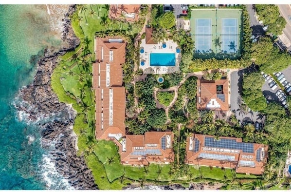 An aerial view of a coastal property with a swimming pool, tennis courts, and solar panels.
