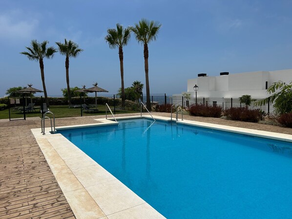 Une piscine avec une terrasse en bois, des palmiers et un bâtiment blanc en arrière-plan.