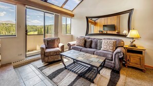 Ein Wohnzimmer mit Sofa, Sessel und Couchtisch, einem an der Wand montierten Fernseher und einem großen Fenster mit Blick auf Wald und Berge.