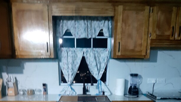 Una cocina con armarios de madera, un lavamanos de acero inoxidable, una ventana con cortina y una estufa.