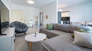 A modern living room with a sofa, a round table, and a chair. There is a kitchen in the background with white cabinets and a stove. - Barbara's sweet home Glyfada by ΜΡS (Glifada)