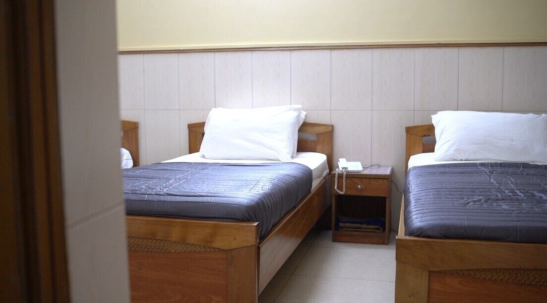 Two wooden beds with white and blue bedding, a wooden nightstand with a phone, and a tiled wall.