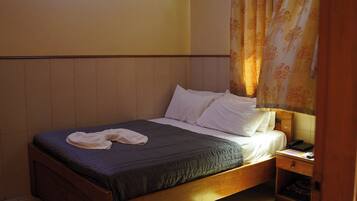 A single bed with a dark green bedspread and white pillows, a wooden headboard, and a nightstand with a phone and papers.