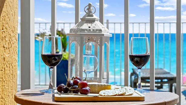 Una mesa con una botella, dos copas de vino tinto, un plato de queso y uvas, y un farol en un balcón con vistas al mar.