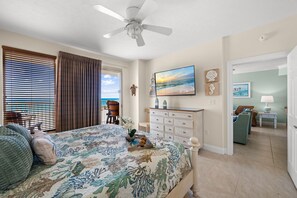 A bedroom with a bed, a ceiling fan, a dresser, a TV, and a view of the ocean.