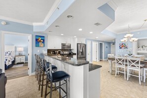 A modern kitchen with white cabinets, a central island with bar stools, stainless steel appliances, and a dining area with white chairs.