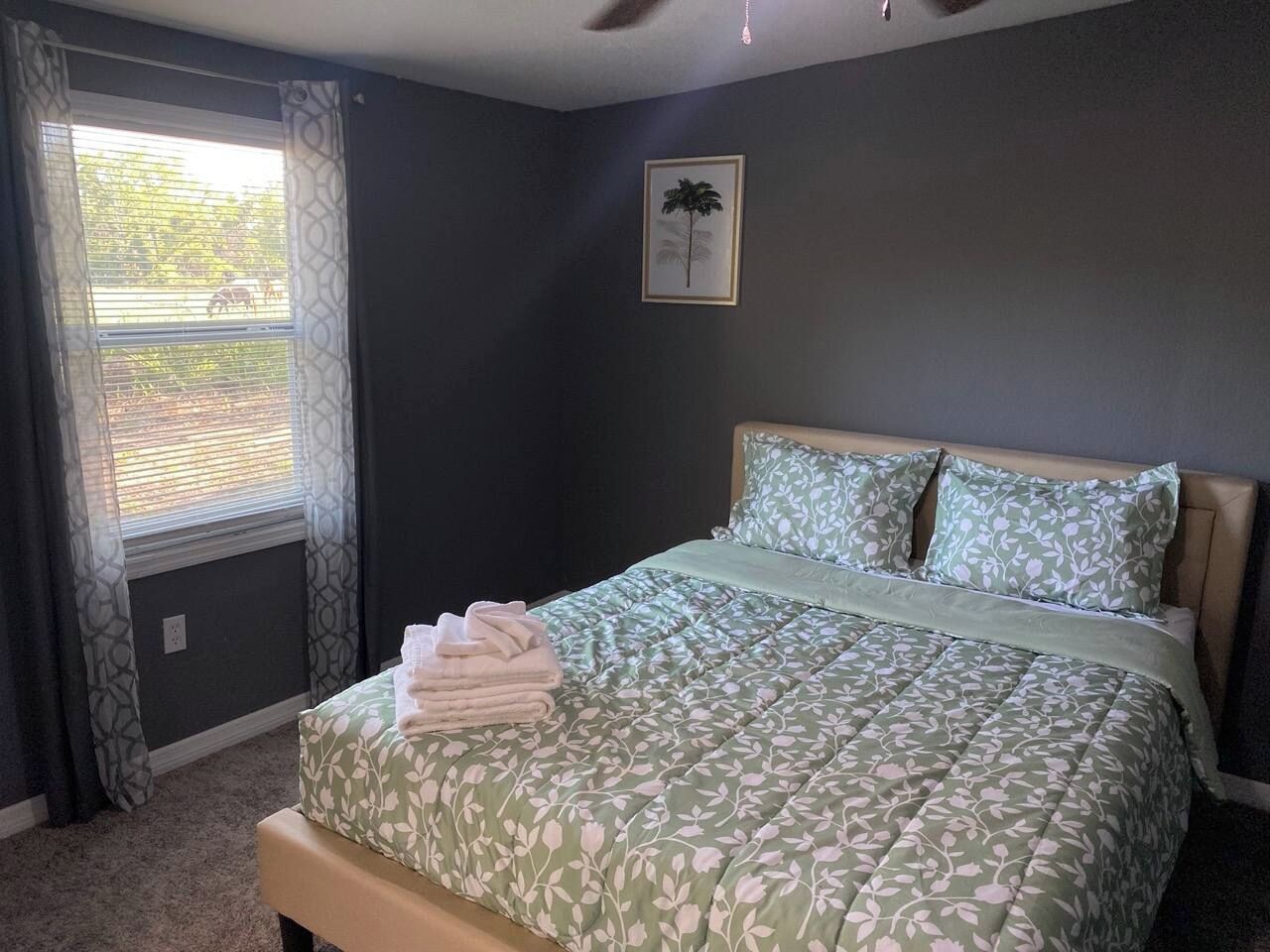 Un dormitorio con cama, ventana con cortinas, un cuadro en la pared y ventilador de techo.