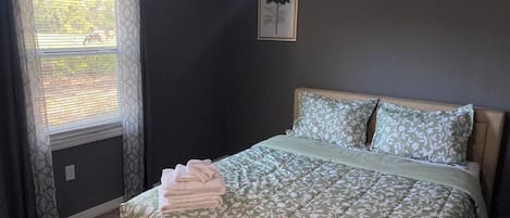 Un dormitorio con cama, ventana con cortinas, un cuadro en la pared y ventilador de techo.