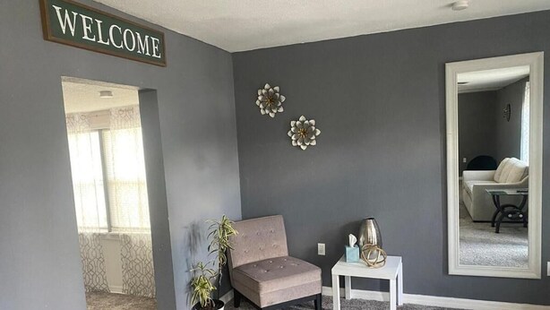 A room with a grey wall, a 'WELCOME' sign, a grey sofa, a small white table, a potted plant, and a mirror reflecting a bed and a chair. - Nature & Comfort: Charming 3BR Palmetto-Ellenton Home, Sleeps 6, WiFi & AC (Palmetto)