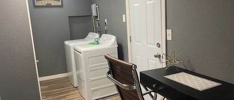Uma lavanderia com máquina de lavar e secadora, uma cadeira e uma escrivaninha com objeto decorativo.