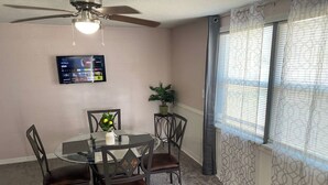 A dining area with a round glass table, four chairs, a ceiling fan, a wall-mounted TV, and a window with patterned curtains. - Palmetto Ellenton | Nature + Smart Comfort (Palmetto)