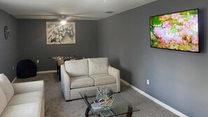A living room with a gray wall, a beige sofa, a glass coffee table, a foosball table, and a ceiling fan. - Nature & Comfort: Charming 3BR Palmetto-Ellenton Home, Sleeps 6, WiFi & AC (Palmetto)