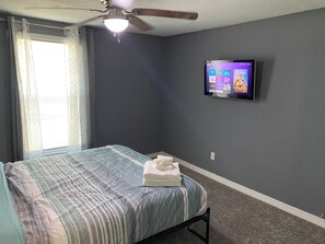 Ein Schlafzimmer mit einem Bett, einem Deckenventilator, einem an der Wand befestigten Fernseher und einem Fenster mit Vorhängen.