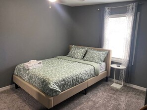 A single bed with a patterned bedspread and pillows, a bedside table with a remote control, and a window with curtains. - Palmetto Ellenton | Nature + Smart Comfort (Palmetto)