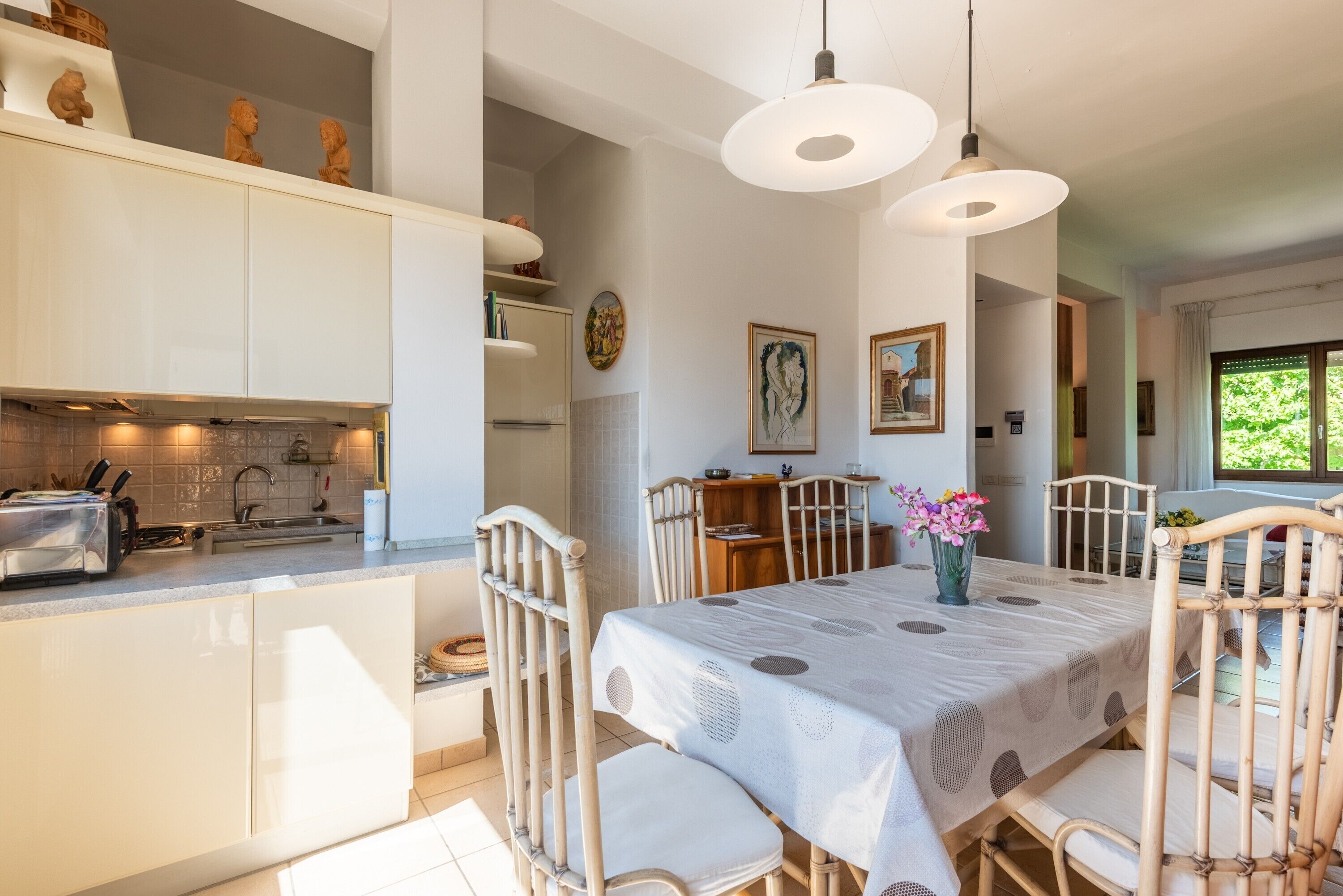 Une cuisine moderne avec des meubles blancs, une table à manger recouverte d’une nappe à motifs et un vase rempli de fleurs.