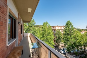 A balcony with a glass table and bench, overlooking a street with parked cars and trees.