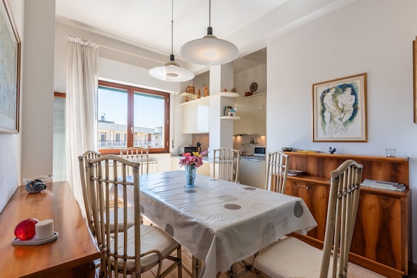 A dining area with a table set for four, chairs, a vase with flowers, and a view of the city through large windows.