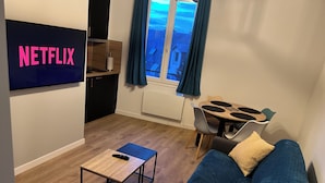 Uma sala de estar com uma TV exibindo 'NETFLIX', uma mesa de centro de madeira, um sofá azul com almofadas e um espaço de jantar com mesa e cadeiras.