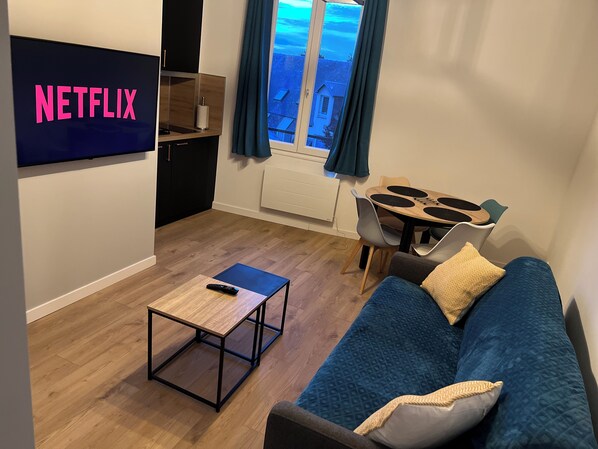 A living room with a TV displaying 'NETFLIX', a wooden coffee table, a blue sofa with cushions, and a dining area with a table and chairs. - Duplex apartment in Honfleur town center, a few minutes from the old port (Honfleur)