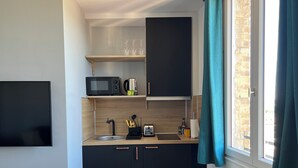 Cozinha compacta com micro-ondas, chaleira e cafeteira na bancada. Há uma janela com cortina e uma TV de tela plana fixada na parede.