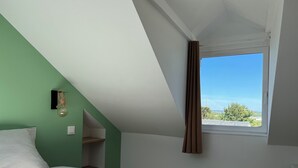 Um quarto com teto inclinado, janela com cortinas, cama com roupa de cama branca e luminária fixada na parede.