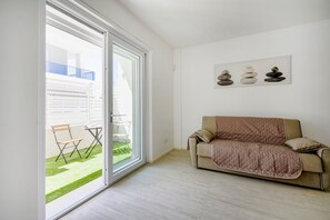 A living room with a sliding glass door leading to a balcony, a beige sofa, and a wall-mounted artwork. - GuestHost - Charming 67 sqm apartment, perfect for 4 people, located on the ground floor of a recently built building.The property is located in a residence with a concierge active all day; outside the house there are gym equipment that can be used freel (Bari)
