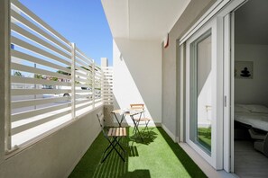 Ein Balkon mit weißer Brüstung, einem kleinen Tisch mit Stühlen und einer Schiebeglastür, die zu einem Schlafzimmer führt.