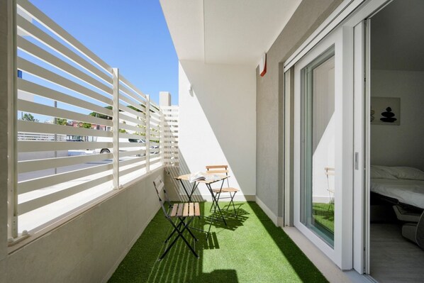 A balcony with a white railing, a small table with chairs, and a sliding glass door leading to a bedroom.