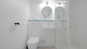 Un baño moderno con inodoro, estante de vidrio con artículos de aseo, lavabo y ducha con mampara de vidrio.