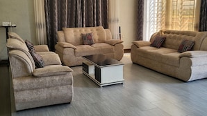 Ein geräumiges Wohnzimmer mit drei Sofas und einem Couchtisch, großen Fenstern mit Vorhängen sowie einem modernen Deckenstil.
