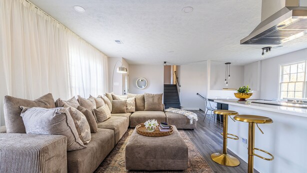 A living room with a sectional sofa, an ottoman, and a coffee table. There is a kitchen area with a bar stool and a window. - Nutmeg (Columbus)