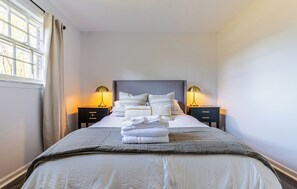 A neatly made bed with pillows and a folded towel, two bedside lamps, and a window with curtains. - Nutmeg (Columbus)