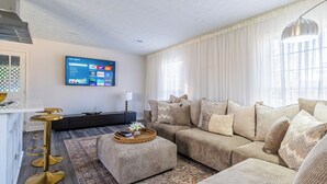 A modern living room with a large flat-screen TV mounted on the wall, a sectional sofa with multiple cushions, a round coffee table with a decorative tray, and a tall floor lamp. - Nutmeg (Columbus)
