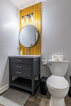 A bathroom with a wooden panel wall, a round mirror, a white toilet, and a black trash bin. - Nutmeg (Columbus)