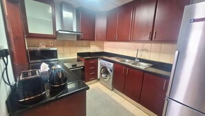 A compact kitchen with dark wooden cabinets, a stainless steel refrigerator, a built-in oven, a washing machine, and a toaster. - Tratoli Hostel in JBR with Sea Views (Dubai)