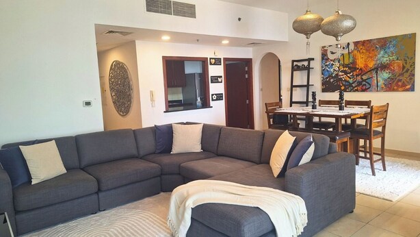 A modern living room with a sectional sofa, a dining area with a table and chairs, and a colorful abstract painting on the wall. - Tratoli Hostel in JBR with Sea Views (Dubai)