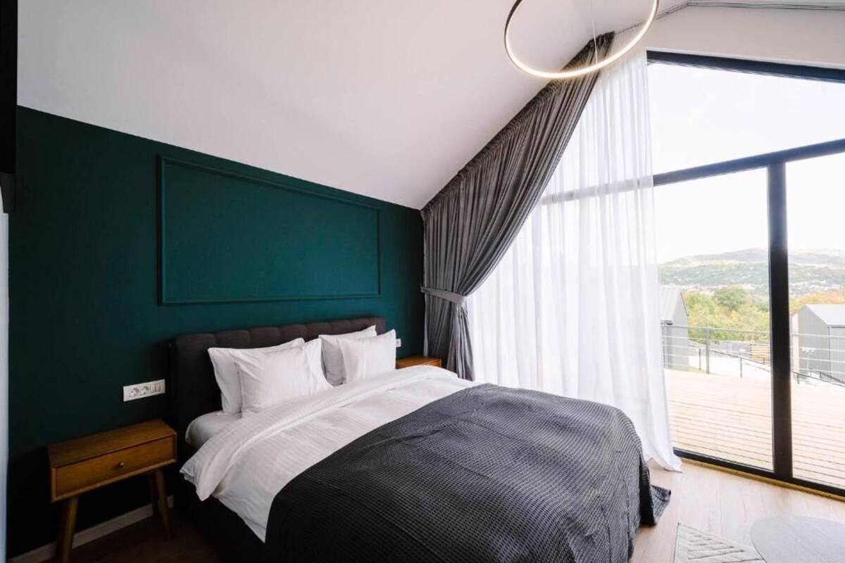 A bedroom with a large bed, a nightstand, a wall-mounted light fixture, and a view of the outdoors through a window.